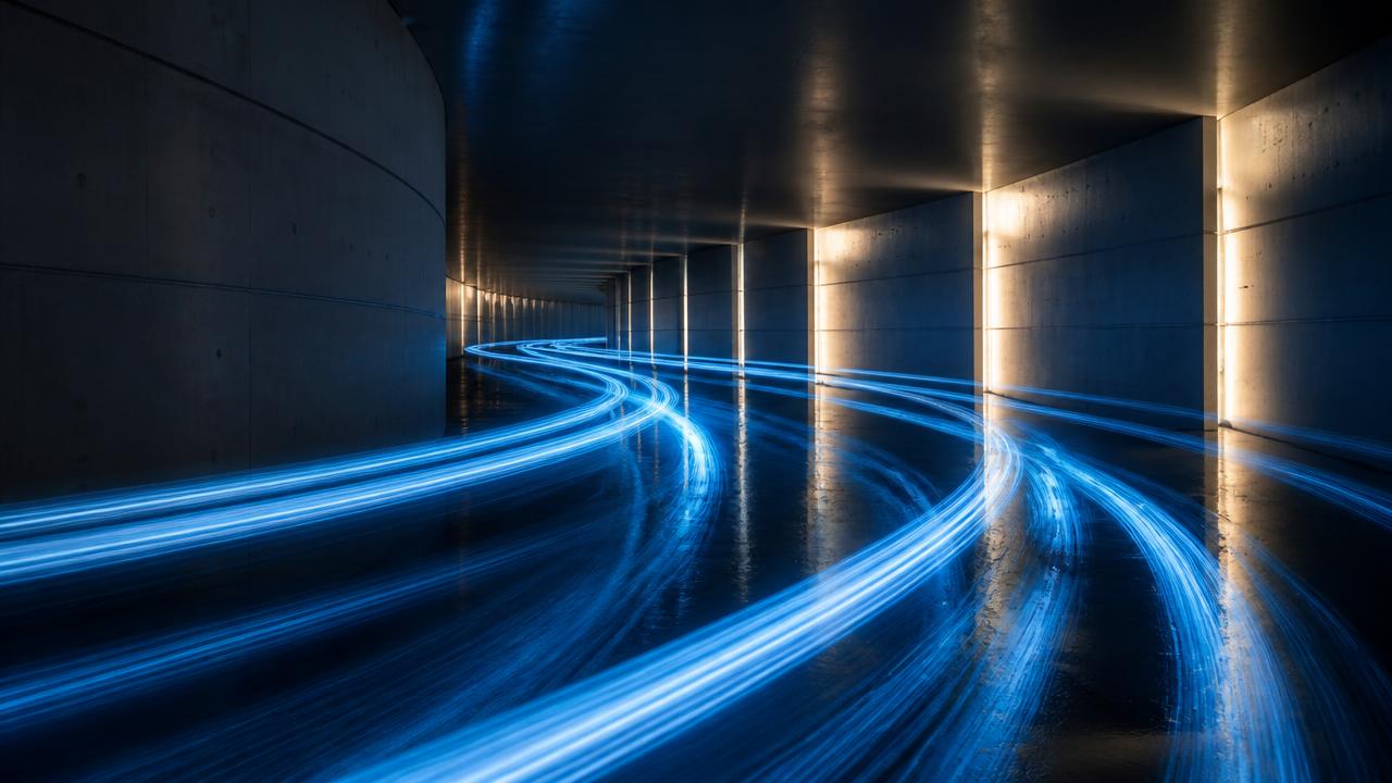 Long-exposure photograph of blue light trails curving through a lit concrete corridor